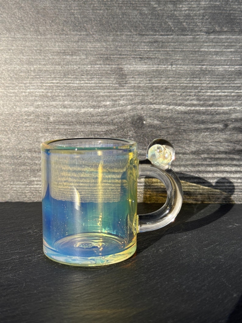 May include: A small, cylindrical glass mug featuring an iridescent finish that shifts between blue and yellow hues. The mug has a clear, curved handle with a small, spherical glass bead accent attached to the top of the handle connection point.