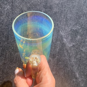 Könnte beinhalten: Schillernder Glasbecher mit einem griffförmigen Totenkopf. Der Becher hat eine zylindrische Form und ein klares, farbenfrohes Finish, das das Licht reflektiert. Der Griff ist ein detailliertes Totenkopfdesign.