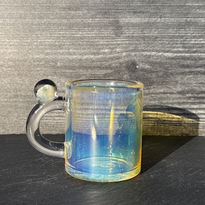 May include: A clear glass mug with an iridescent finish that reflects blue and yellow tones. The mug features a curved handle topped with a small, spherical glass bead accent. The cylindrical body is transparent with a smooth, polished surface.