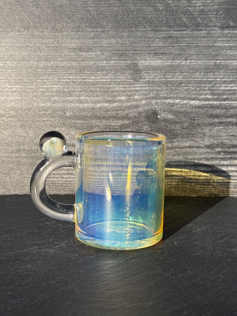 May include: A clear glass mug with an iridescent finish that reflects blue and yellow tones. The mug features a curved handle topped with a small, spherical glass bead accent. The cylindrical body is transparent with a smooth, polished surface.