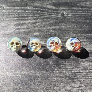 May include: Four glass marbles with a skull design inside. The marbles are clear and have a iridescent finish. The skulls are white and have a detailed design.