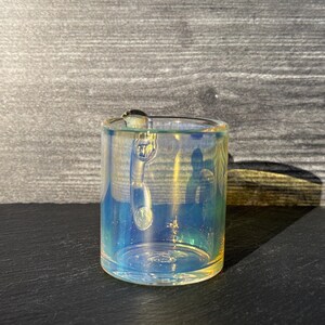 May include: A cylindrical glass mug featuring an iridescent finish that reflects shades of blue, yellow, and green. The mug has a clear glass handle attached to the side and a smooth, transparent rim, showcasing a reflective surface quality.