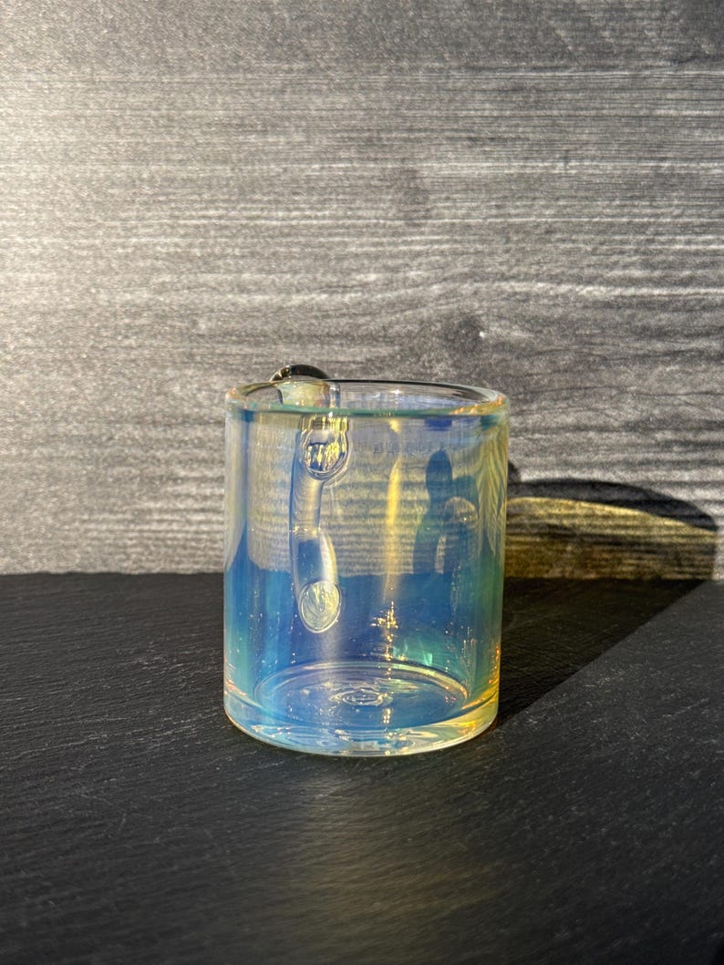 May include: A cylindrical glass mug featuring an iridescent finish that reflects shades of blue, yellow, and green. The mug has a clear glass handle attached to the side and a smooth, transparent rim, showcasing a reflective surface quality.