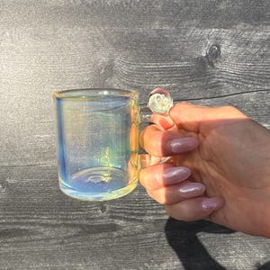 May include: A small, clear glass mug featuring an iridescent finish that reflects shades of blue, yellow, and pink. The mug has a cylindrical body and a small, rounded glass handle attached to the side, creating a shimmering, multi-colored effect.