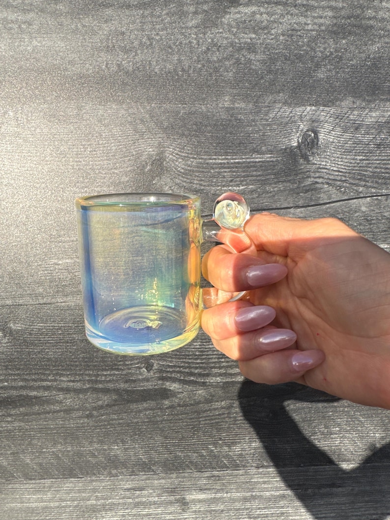 May include: A small, clear glass mug featuring an iridescent finish that reflects shades of blue, yellow, and pink. The mug has a cylindrical body and a small, rounded glass handle attached to the side, creating a shimmering, multi-colored effect.