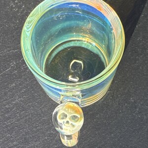 May include: A clear glass mug with an iridescent finish that reflects blue and yellow tones. The mug features a circular handle with a three-dimensional skull detail embedded within the glass at the top of the handle, viewed from above.