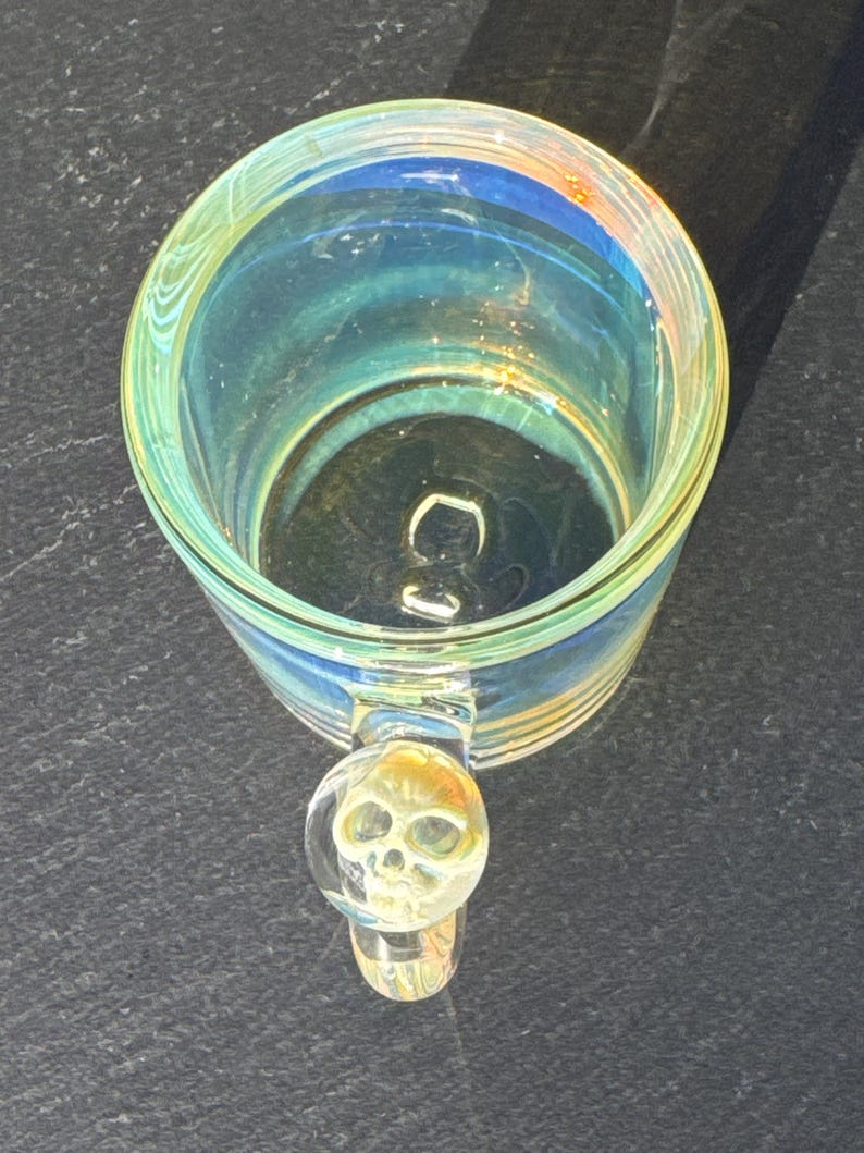 May include: A clear glass mug with an iridescent finish that reflects blue and yellow tones. The mug features a circular handle with a three-dimensional skull detail embedded within the glass at the top of the handle, viewed from above.