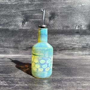 Könnte beinhalten: Eine dekorative Glasflasche mit einem Ausgießer aus Edelstahl. Die Flasche ist eine Mischung aus Gelb und Blau mit einem Muster aus Kreisen. Die Flasche ist zum Ausgießen von Flüssigkeiten bestimmt.