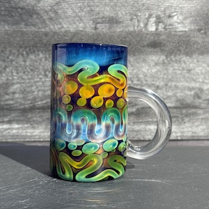 Könnte beinhalten: Ein zylindrischer Glasbecher mit einem durchsichtigen Henkel. Der Becher hat ein farbenfrohes Design mit blauen, grünen, gelben und orangenen Wirbeln und Punkten. Die Oberseite des Bechers ist tiefblau.