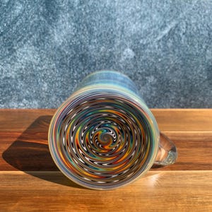 Pode incluir: Uma caneca de vidro com um design em espiral colorido na parte inferior. A caneca tem uma alça transparente e uma forma cilíndrica. O padrão em espiral inclui tons de azul, laranja, amarelo e preto. A caneca está em uma superfície de madeira.