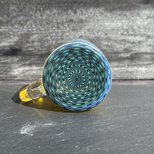 Op de afbeelding: Een glazen mok met een gele handgreep en een blauw en blauwgroen spiraalontwerp aan de onderkant. De mok is gemaakt van helder glas en staat op een donkere ondergrond.