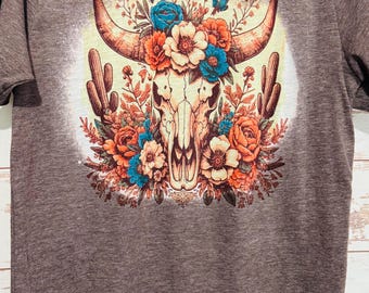 May include: A brown t-shirt with a floral skull design. The skull is surrounded by flowers in shades of orange, blue, and white. The background is a light yellow.