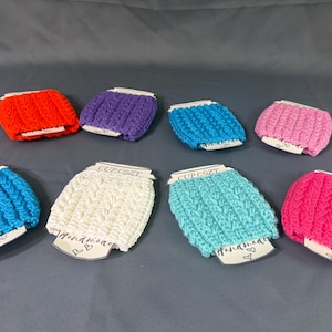 May include: A collection of eight crocheted cup cozies in various solid colors including red, purple, blue, pink, white, and turquoise. Each cozy features a textured cable knit pattern and is attached to a white paper tag labeled CUP COZY.