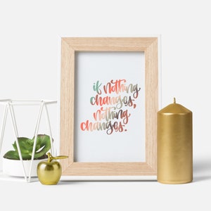 May include: A framed print with the text "if nothing changes, nothing changes" in colourful script. A gold candle, a white geometric planter with a succulent, and a gold apple figurine are also present.