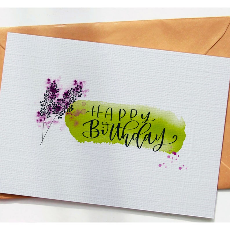 May include: A white card with a green watercolor wash and the text "Happy Birthday" in black calligraphy. There are purple watercolor flowers in the top left corner.