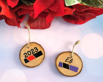 May include: Two wooden ornaments with twine hangers. One ornament displays the year "2023" above a black, red, and black design. The other ornament has the name "Eli" above a blue, black, and purple design. Red flowers are in the background.