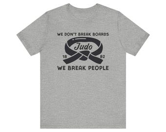 May include: A gray t-shirt with a black graphic. The graphic features a Judo belt design with the word "Judo" in the center, along with the text "We don't break boards, we break people" and the numbers "18" and "82".