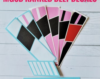 May include: A set of six colorful belt decals for martial arts, featuring black, pink, red, and white stripes. The decals are arranged in a fan shape and have the text "MGSD RANKED BELT DECALS" and "MEANGIRLSELFDENSE.COM" printed on them.