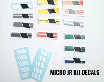 May include: A set of decals for a Micro JR BJJ uniform. The decals are in various colors, including yellow, red, green, blue, and gray. The decals feature a black stripe and a black rectangle. There are also two sheets of white decals with a repeating pattern of vertical lines.