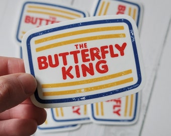 May include: A white sticker with a blue border and yellow stripes. The text "THE BUTTERFLY KING" is in red letters.