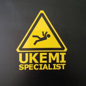 Puede incluir: Una señal de advertencia amarilla con una figura de palo cayendo dentro. El texto debajo de la señal dice "UKEMI SPECIALIST".