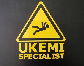May include: A yellow warning sign with a stick figure falling down inside. The text below the sign reads "UKEMI SPECIALIST".