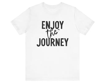 May include: White t-shirt with the phrase "ENJOY the JOURNEY" printed in black. The word "the" is in a handwritten style, with a small pencil graphic. The shirt has short sleeves and a crew neck.