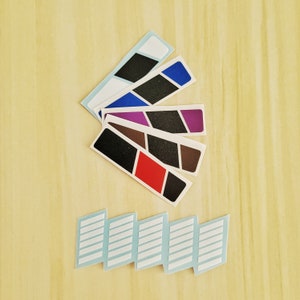 Op de afbeelding: Vijf verschillende gekleurde plakstrips met een diagonaal patroon. De strips zijn in een waaier vorm gerangschikt. Onder de strips bevinden zich zes witte rechthoekige plakstrips.