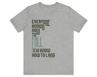 May include: A heather gray t-shirt with the text "EVERYONE KNOWS HOW TO FALL, EW KNOW HOW TO LAND" in black and green lettering. The shirt is a short-sleeved crew neck.