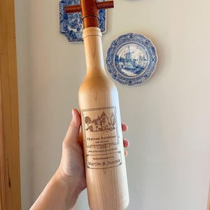 May include: A wooden wine bottle-shaped stopper with a dark brown tap. The bottle has a light wood finish and features a label with the text "Chateau Bordeaux 1982 Vintage" and the names "Martin & Jeannie". Decorative plates are in the background.