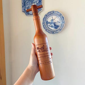 May include: A wooden wine bottle-shaped bottle stopper with a cross-shaped handle. The bottle stopper has a label with the text "Chateau Bordeaux 1982 Vintage" and "Bottled Especially For Martin & Jeannie."