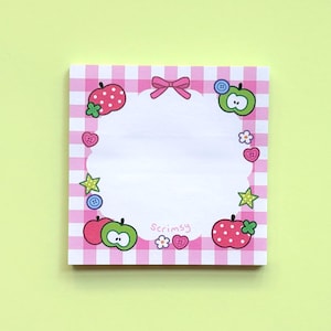 May include: A square notepad with a pink and white gingham border. The center features a white scalloped area decorated with illustrations of red and green apples, small buttons, stars, flowers, and a pink bow. The text Scrimsy is printed at the bottom.