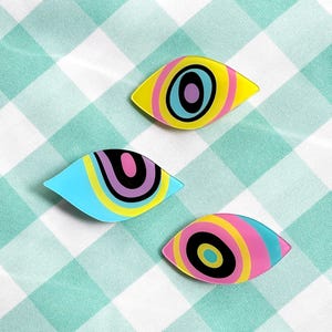 以下が含まれることがあります： 鮮やかな色の同心円が描かれた、目のような形のピンバッジ3点。上のピンは黄色、水色、紫。左のピンは水色、黄色、黒、ピンク。下のピンはピンク、黄色、水色、黒で構成されています。