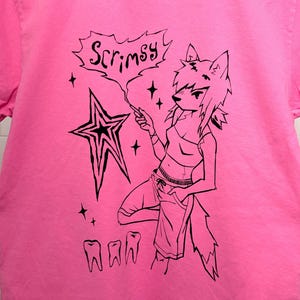 May include: A bright pink t-shirt featuring a black graphic design. The design includes a stylized wolf-like character, a star, the word "Scrimsy" in a speech bubble, and three teeth. The shirt is hanging on a white hanger.
