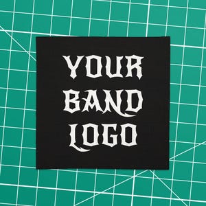 Könnte beinhalten: Ein schwarzer quadratischer Aufnäher mit dem weißen Schriftzug "YOUR BAND LOGO" in gotischer Schrift. Der Aufnäher liegt auf einer grünen Schneidematte mit weißen Gitterlinien. Ideal zum Anpassen von Kleidung oder Accessoires.