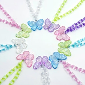 May include: A selection of butterfly necklaces in various colours. Each necklace has a translucent butterfly pendant in shades of pink, blue, purple, green, and clear, attached to a matching chain. The necklaces are arranged in a circular pattern.