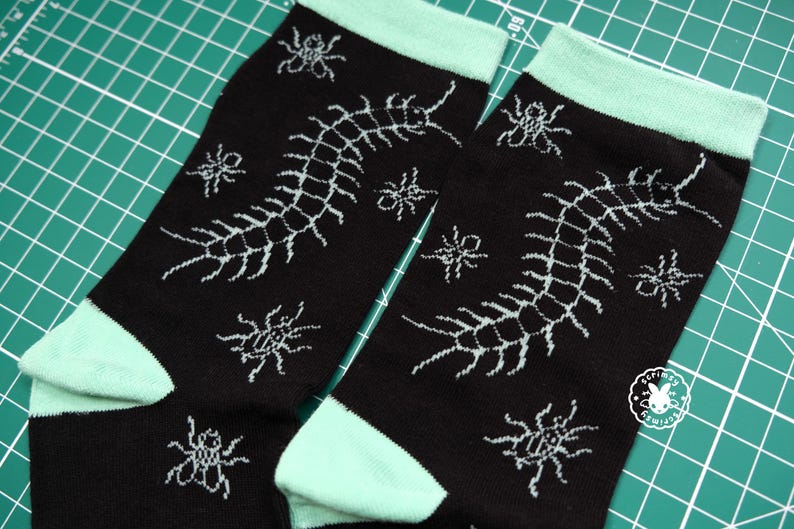 May include: A pair of black socks with a mint green trim. The socks feature a white graphic print of a centipede and spiders.