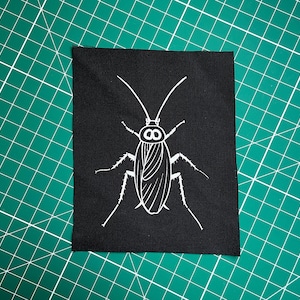 Puede incluir: Parche de tela negro con un contorno blanco de una cucaracha. La cucaracha tiene dos ojos grandes y antenas largas.