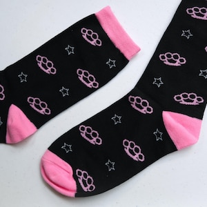 Pu&ograve; includere: Un paio di calzini neri con un motivo a forma di nocche rosa. I calzini hanno un bordo rosa intorno alla parte superiore e al tallone.