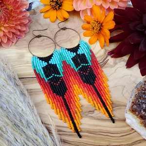 Pode incluir: Brincos de contas feitos à mão com um design vibrante. Os brincos apresentam contas turquesa, pretas, vermelhas e laranja, criando um padrão colorido. Eles têm aros cor de cobre e franjas longas. Os brincos são exibidos com flores e um cristal.