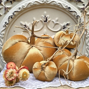 Puede incluir: Un conjunto de cuatro calabazas de terciopelo dorado con tallos secos realistas y un pequeño grupo de flores de paja secas con pétalos rosas y amarillos. Las calabazas son suaves, redondeadas y presentan profundos segmentos verticales de tela.