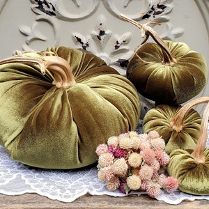 May include: Olive green velvet pumpkins of varying sizes with realistic stems. A small bouquet of pink and beige dried flowers sits near the pumpkins. The pumpkins are arranged on a white lace doily.