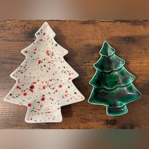 Puede incluir: Dos platos de cerámica con forma de árbol de Navidad. El plato más grande es blanco con salpicaduras de pintura roja y verde. El plato más pequeño presenta un esmalte verde oscuro brillante y sólido con sutiles sombras negras en los bordes.
