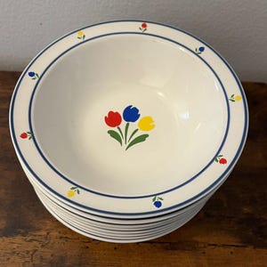 May include: A stack of white ceramic bowls featuring a central motif of red, blue, and yellow tulips with green leaves. The rim of each bowl is decorated with a thin blue circular line and small, alternating red, blue, and yellow tulip icons.
