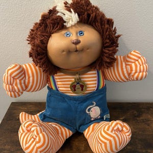 May include: A plush doll with a vinyl face, blue eyes, and brown yarn hair with a white streak. It wears an orange and white striped long-sleeve shirt, blue corduroy shorts with an elephant patch, and rainbow suspenders. A gold medallion hangs from its neck.