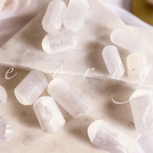 May include: A collection of white selenite crystal wands on a white marble surface. The crystals are smooth and polished, with a translucent appearance.