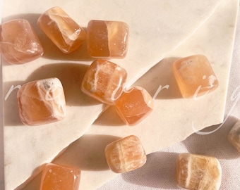 May include: A collection of orange calcite tumbled stones, with a variety of patterns and textures. The stones are arranged on a white surface.