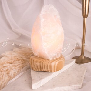 May include: A white Himalayan salt lamp with a wooden base. The lamp is lit and casting a warm glow. The lamp is sitting on a white marble surface.