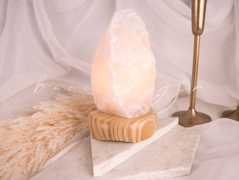 May include: A white Himalayan salt lamp with a wooden base. The lamp is lit and casting a warm glow. The lamp is sitting on a white marble surface.