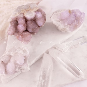 May include: An assortment of crystals in shades of lavender and clear quartz are arranged on a white marble surface. The crystals include clusters and points, with the text "bare origin" in a script font.